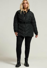 Schwarze Daunenjacke mit tailliertem Gürtel, Reißverschluss und Zickzackmuster. Kombiniert mit schwarzen, eng anliegenden Hosen und klobigen schwarzen Stiefeln.