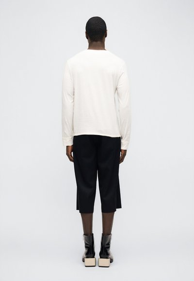 Άτομο στέκεται με την πλάτη γυρισμένη φορώντας λευκό πουκάμισο με μακριά μανίκια, μαύρο φαρδύ παντελόνι cropped και μαύρα μποτάκια αστραγάλου με χοντρές γόβες.