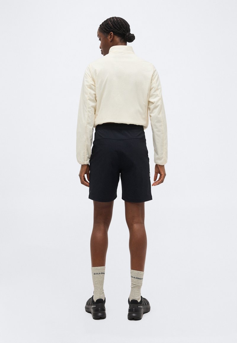 Personne aux cheveux tressés en chignon portant une veste crème, un short noir, des chaussettes beiges et des baskets noires, se tenant de dos sur un fond uni.