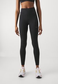 Hochgeschnittene schwarze Leggings mit glatter Textur, ausgestattet mit einer Seiten Tasche und einem dezenten Logo in der Nähe der Taille. Kombiniert mit weißen Sportschuhen.