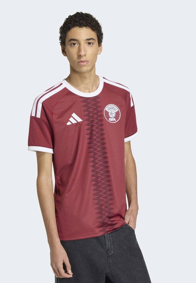 Vinröd fotbollströja med vita detaljer, rund halsringning och zickzackmönster i mitten. Har en emblem och tre-randigt mönster på axlarna.