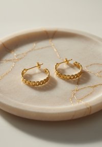 Guldörhängen med en flätad design, visade på en rund marmorplatta med en ljus färg och guldådring.