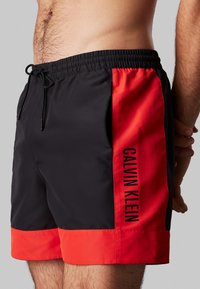 Costumi da bagno da uomo in nero e rosso. Realizzati in tessuto morbido, con vita elastica, cordino e stampa del logo laterale.