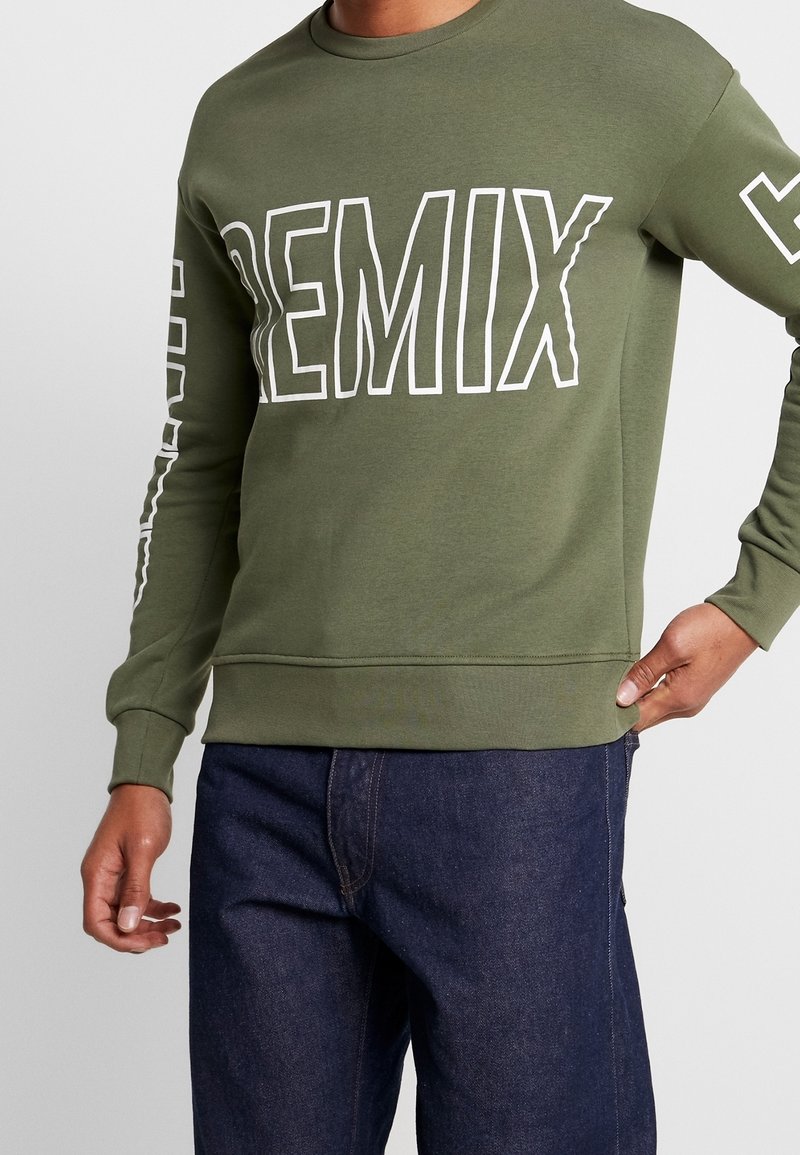 Sudadera de color verde oliva con el texto blanco "REMIX" en el frente y a lo largo de la manga izquierda. Cuenta con un cuello redondo y dobladillos ribeteados en el bajo y los puños.