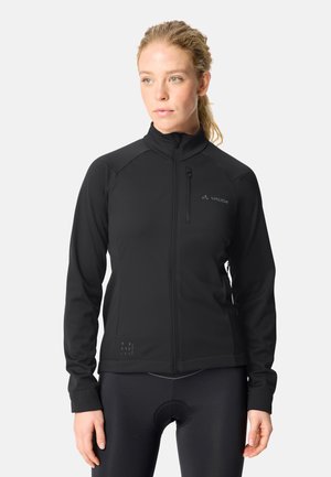 Vrouw met licht haar in een paardenstaart, draagt een zwarte fietsjas met rits en zwarte gewatteerde fietsbroek, kijkt iets opzij.