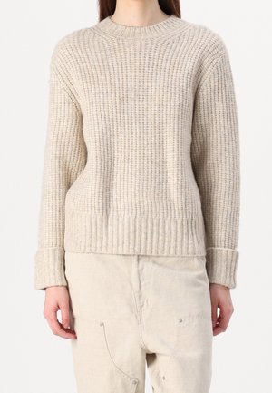 Femme portant un pull en maille côtelée beige à manches longues et un pantalon décontracté blanc cassé avec coutures visibles et poches.