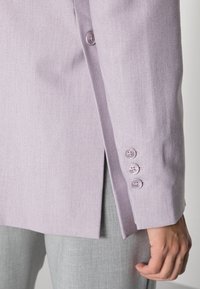 Světle fialový blazer s texturovanou látkou, s bočním rozparkem a třemi odpovídajícími knoflíky na manžetě, ve spojení se světle šedými kalhotami.