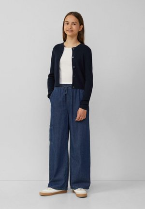 Navy cardigan over a white t-shirt, paired with wide-leg denim trousers featuring a pocket. White sneakers with a gum sole.