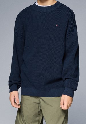 Garçon portant un pull en maille texturée bleu marine foncé avec un petit logo sur la poitrine, associé à un pantalon vert olive sur fond gris uni.