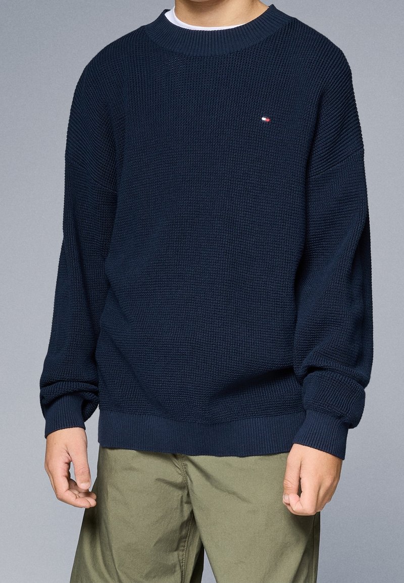 Garçon portant un pull en maille texturée bleu marine foncé avec un petit logo sur la poitrine, associé à un pantalon vert olive sur fond gris uni.