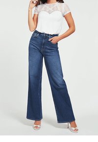 Kvinde iført højtaljede, brede bukser i blå denim, hvid spidse ærmer top af blonder og hvide stilethæl sandaler, der står med hånden i lommen.