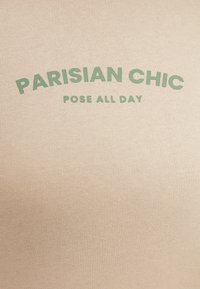 Top de algodón beige con texto verde que dice "PARISIAN CHIC" y "POSE ALL DAY", con una textura suave y diseño de letras curvadas.