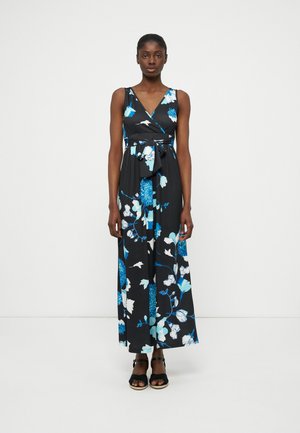 Γυναίκα που φοράει μαύρο αμάνικο μάξι φόρεμα με μπλε και λευκά λουλούδια και ζώνη στη μέση, στέκεται μπροστά σε ένα απλό ανοιχτόχρωμο φόντο.