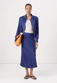Giubbotto bomber blu con vita sagomata, abbinato a una maglietta beige e una gonna midi blu. La modella tiene una borsa arancione e indossa sandali neri.