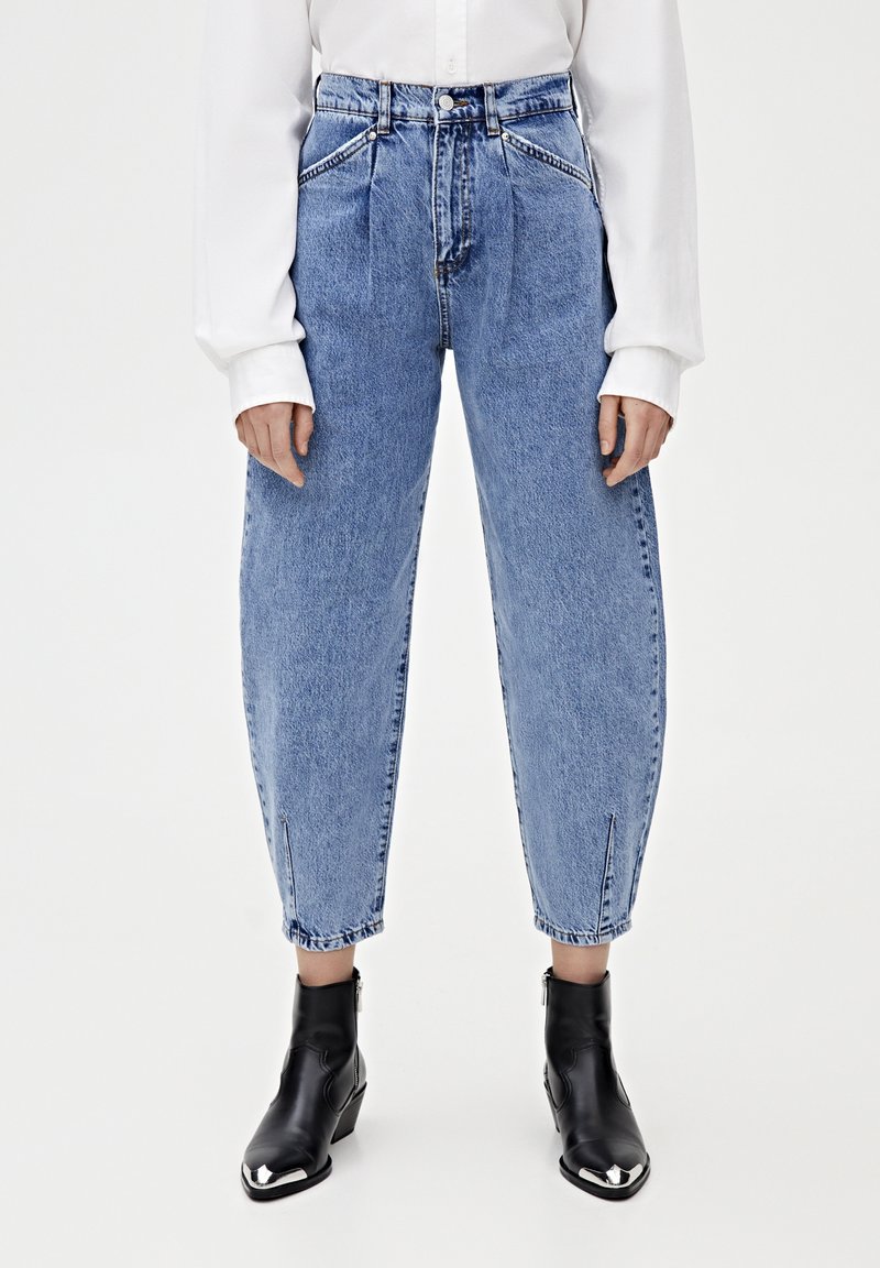 Femme portant un jean bleu clair plissé taille haute avec des fentes aux chevilles, associé à des bottines noires à bout pointu et une chemise blanche à manches longues.