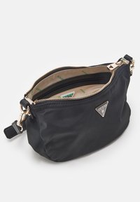 Sort Guess håndleddtaske med guld lynlås, beige indvendig foret med logoer og en trekantet metal-logo plade på forsiden.