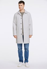 Impermeabile grigio chiaro con colletto, sei bottoni, e maniche lunghe, indossato sopra un maglione verde a maglia e jeans blu. Sneakers bianche.
