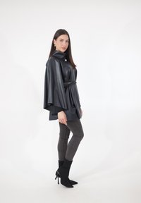 Schwarzes Cape-Oberteil aus Kunstleder mit hohem Kragen, Taillengürtel, drapierten Ärmeln, kombiniert mit dunklen Slim-Jeans und schwarzen Stiefeletten.