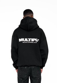 Černá mikina s kapucí z měkké tkaniny má na zadní straně velké bílé logo "MULTIPLY" a šňůrku na stažení kapuce. Nenáročně kombinována s tmavými džíny.