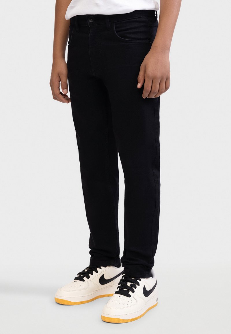 Blugi negri din denim cu croială slim, având un design cu cinci buzunare și detalii subtile de cusătură, asortați cu adidași albi cu accente negre.