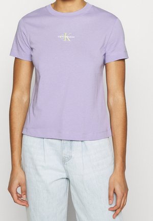 Persoon draagt een lila Calvin Klein Jeans t-shirt en lichtblauwe high-waisted jeans en staat tegen een effen witte achtergrond.
