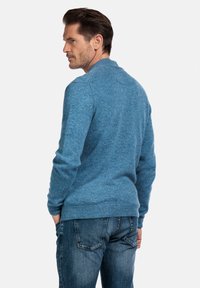 Blauwe gebreide trui met een gestructureerde afwerking, geribbelde manchetten en tailleband, gecombineerd met donkere spijkerbroek, gezien van achteren.