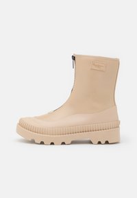 Bota de goma beige a media pantorrilla con suela gruesa acanalada, cierre delantero y pequeño parche de marca en el lado exterior.