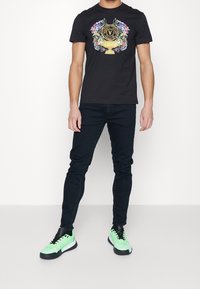 Musta t-paita, jossa on värikäs graafinen kuvasto, jossa on maljakko ja teksti, yhdistettynä tummiin farkkuihin ja neonvihreisiin urheilujalkineisiin.