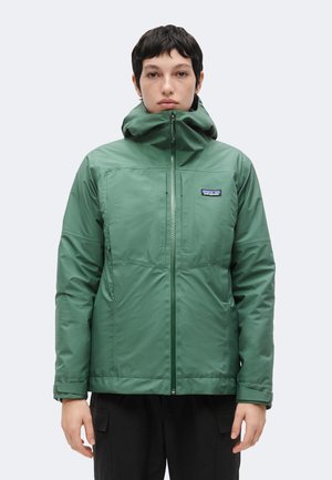 Person mit kurzem dunklem Haar trägt eine grüne Kapuzenjacke mit Reißverschluss und schwarze Hose, steht vor einem einfarbigen hellen Hintergrund.