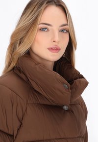 Cappotto imbottito marrone con colletto alto e cuciture prominenti. Presenta bottoni metallici vicino al colletto. Il materiale ha una texture liscia e lucida.