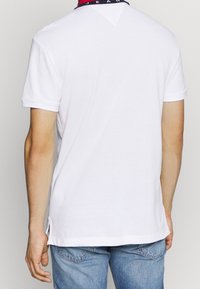 Bílá polo košile s krátkým rukávem zobrazená zezadu, s kontrastním červeným a námořnickým límcem s černými detaily a bočními štěrbinami.