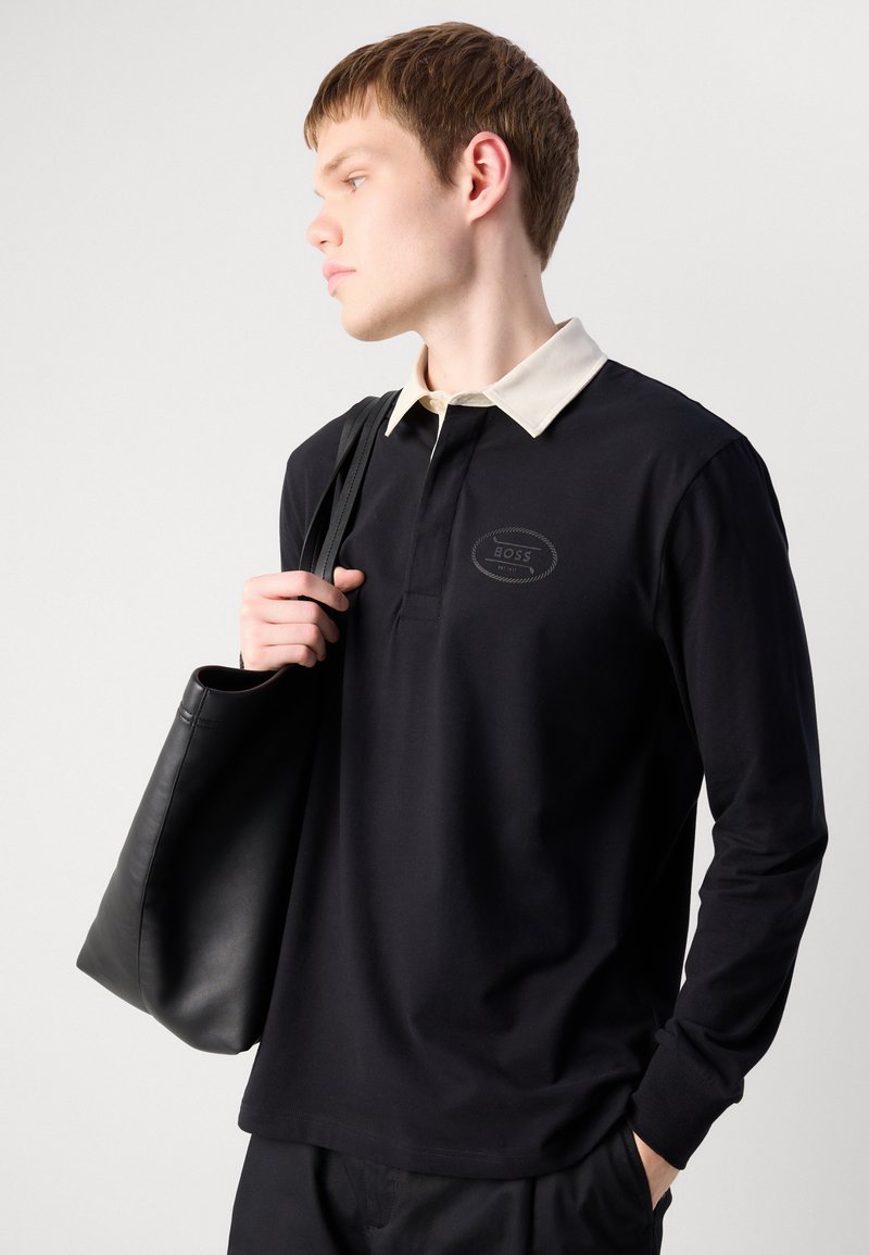 Zwarte poloshirt met lange mouwen, voorzien van een crèmekleurige kraag en een knoopsluiting aan de voorkant. Heeft een subtiele merklogo op de borst. Vergezeld van een zwarte tote bag.