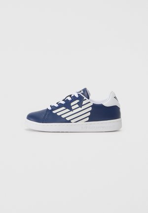 Baskets bleu marine avec lacets blancs, semelle blanche et grand logo d’aile blanche sur le côté sur fond uni.