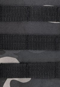 Gros plan sur des sangles en nylon noir cousues sur un tissu camouflage gris foncé.