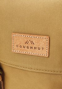 Beige kanvasbag med teksturert overflate, preget lærmerke med "DOUGHNUT" og kontrasterende hvit søm.