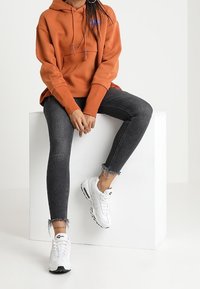 En person i en orange hoodie, slitna svarta tajta jeans och vita sneakers sitter på en vit kub mot en enkel bakgrund.