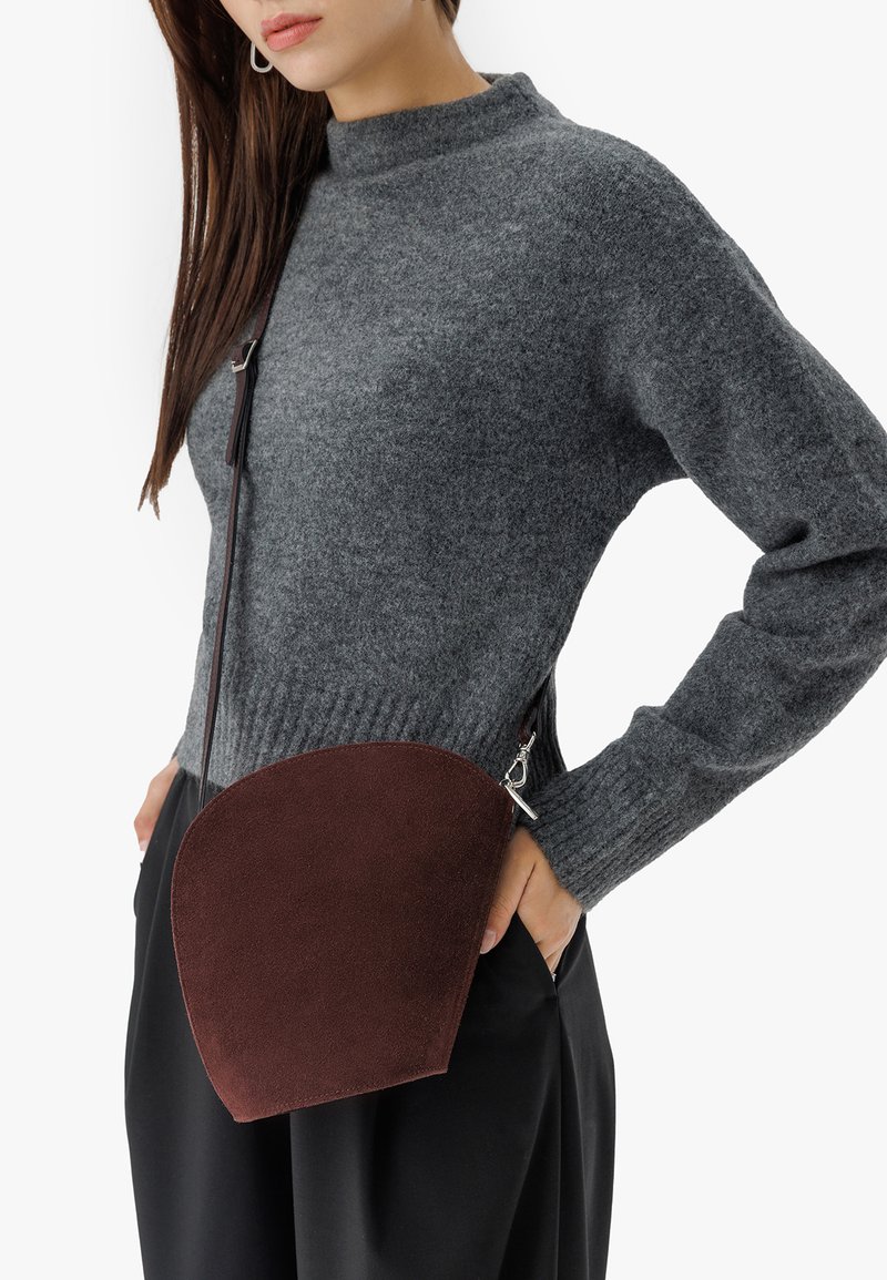 Brūns zamšādas krustsoma ar pusapaļo formu, sudraba piederumiem un regulējamu siksniņu, uzvilkta kopā ar pelēku džemperi un melnām biksēm.