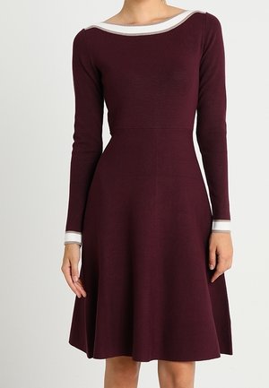 Femme portant une robe bordeaux à manches longues avec une bordure blanche au niveau de l'encolure et des poignets, debout devant un fond uni.