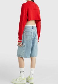 Červený zkrácený pletený svetr, světle modré džínové šortky s boční kapsou, bílá tričko pod tím, bílé a zelené tenisky, žebrované ponožky.