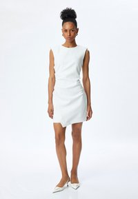 Witte geribbelde mouwloze jurk met een asymmetrische zoom. Gecombineerd met witte spitse hakken. Zachte textuur en aansluitende silhouet.