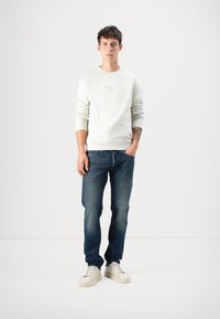Young man standing with hand in pocket wearing white sweatshirt, blue jeans, off-white sneakers, and visible forearm tattoo.