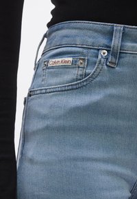 Svijetloplave traperice klasičnog dizajna s pet džepova, s oznakom Calvin Klein, srebrnim metalnim detaljima i narančastim šavovima.