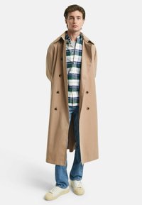 Gabardina beige con grandes botones sobre una camisa de cuadros verde y azul, combinada con jeans azul claro y zapatillas blancas.