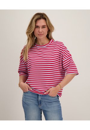 Vrouw met lang blond haar die een rood-wit gestreept shirt en blauwe spijkerbroek draagt, staat met de handen in de zakken tegen een effen achtergrond.