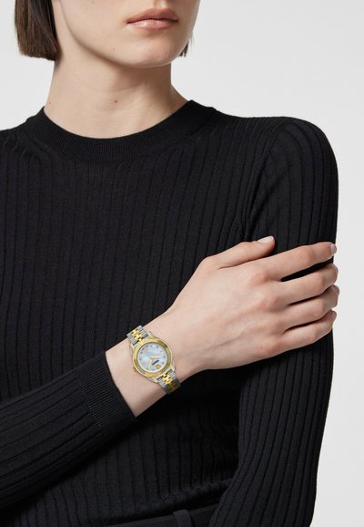 Reloj de metal dorado y plateado con una cara redonda, esfera azul clara y bisel texturizado. Lleva puesto en una muñeca con un suéter negro de canalé.