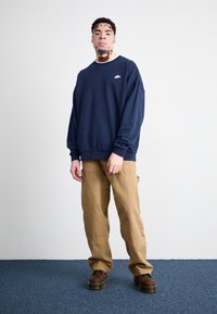 Sudadera azul marino con cuello redondo y logo, combinada con pantalones cargo caqui y botas marrones. Fondo liso con alfombra azul.