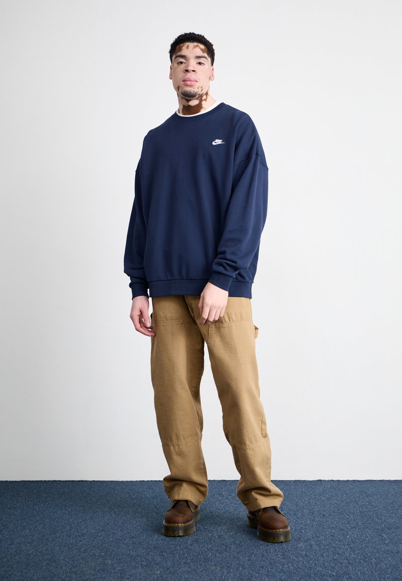 Sweatshirt bleu marine à col rond avec logo, associé à un pantalon cargo beige et des bottes marron. Fond uni avec un tapis bleu.