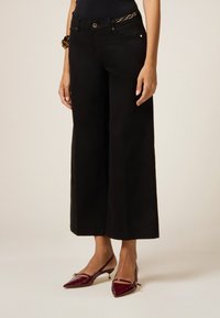 Pantalones de pierna ancha negros con una textura suave, presentan dos bolsillos frontales y un acento de cadena dorada en la cintura, combinados con tacones de charol color burdeos.