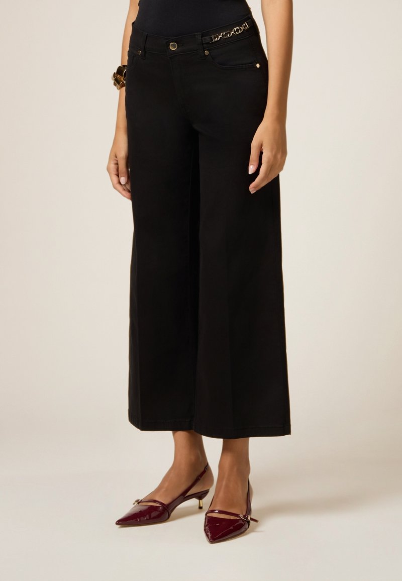 Pantalones de pierna ancha negros con una textura suave, presentan dos bolsillos frontales y un acento de cadena dorada en la cintura, combinados con tacones de charol color burdeos.