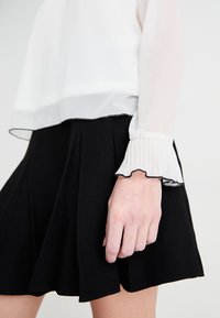 Blouse blanche en voile avec un bord froncé noir aux poignets, associée à une jupe plissée noire, mettant en valeur des textures douces et des couleurs contrastées.